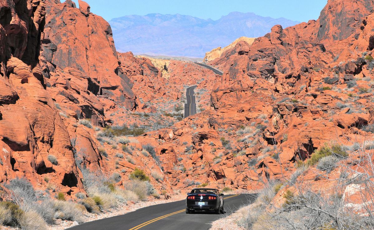 A dark convertible drives along a winding paved road through red sandstone rock formations, with distant blue mountains under a clear sky.