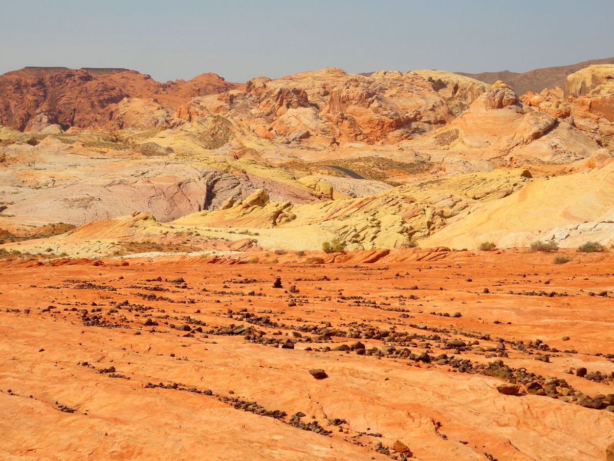 Wide desert view of orange, yellow and pale sandstone hills and rock slabs scattered with dark stones under a hazy blue sky.