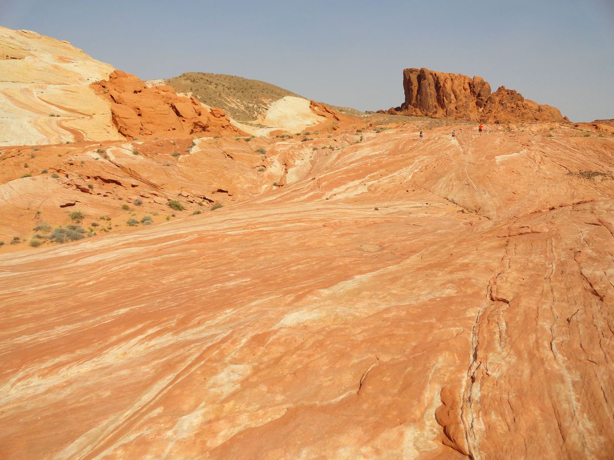 Wide desert landscape of smooth orange and cream striped sandstone with a rugged red rock formation on the horizon under a pale blue sky.